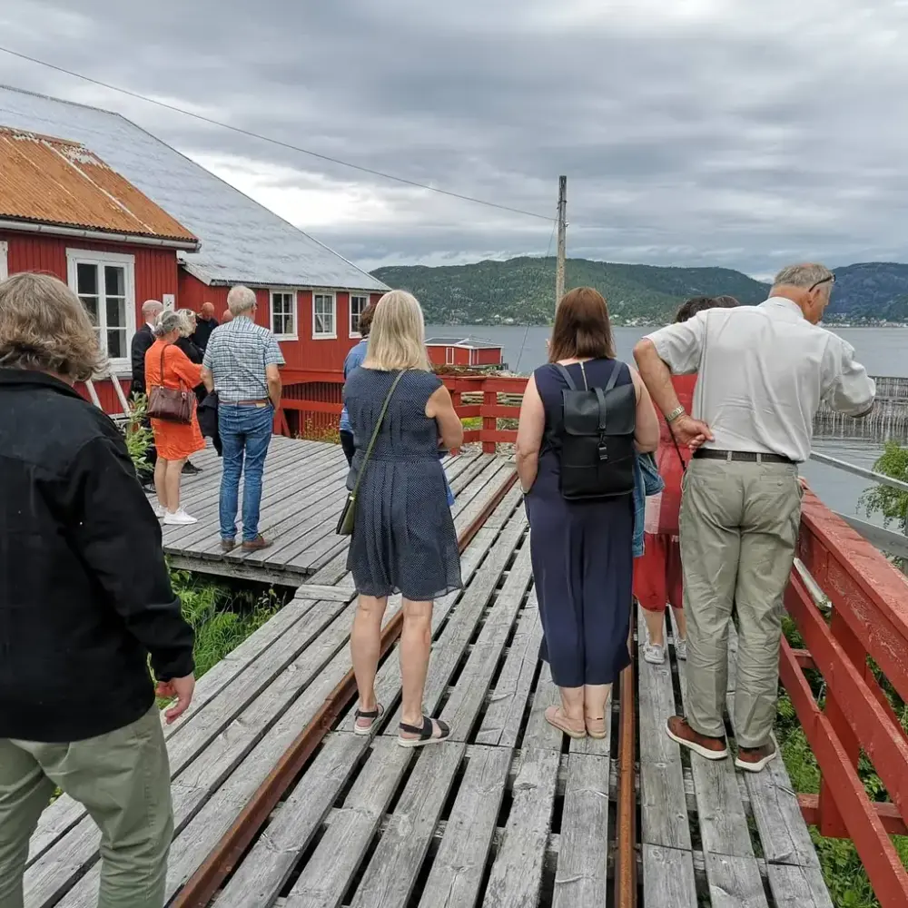 En gruppe mennesker står utenfor et rødt trebygg og ser ned mot vannet og en tømmerbom. Foto.