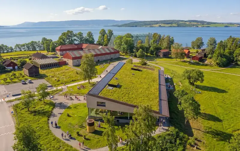 Dronefoto av Anno Domkirkeodden historiesenter med grønt tak, omgitt av grønne trær og kulturlandskap ved Mjøsa.