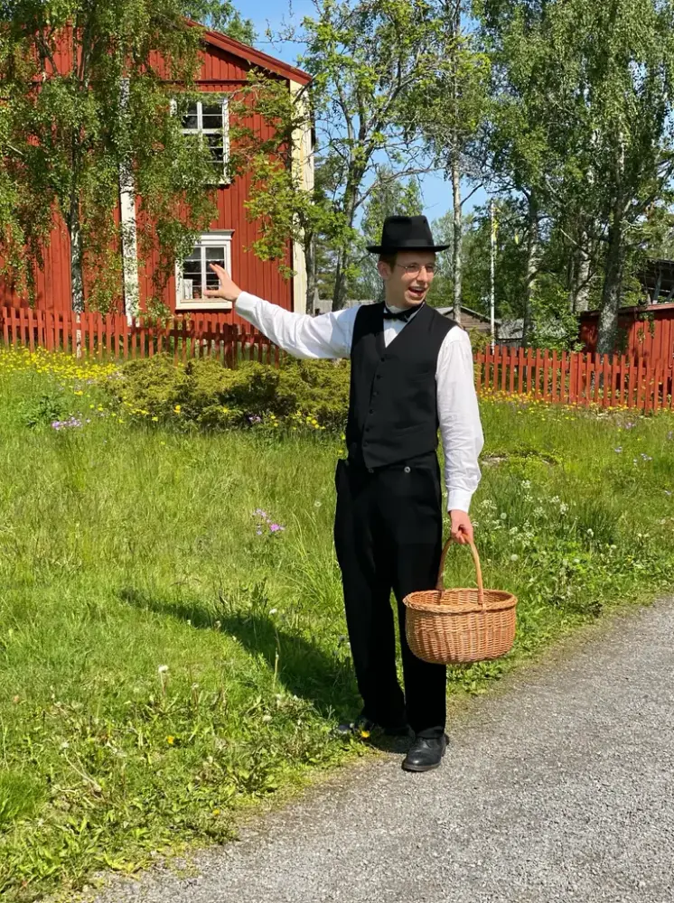 En man med svarta kläder och svart hatt står med en korg i grönt gräs och ett rött hus i bakgrunden.