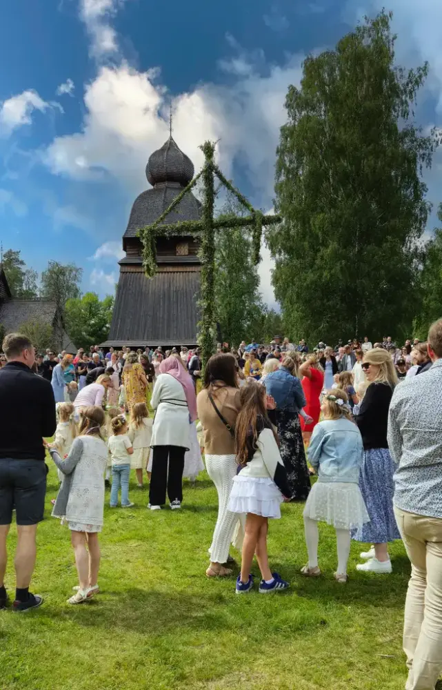 En folksamling runt en midsommarstång med en mörkfärgad byggnad i bakgrunden.