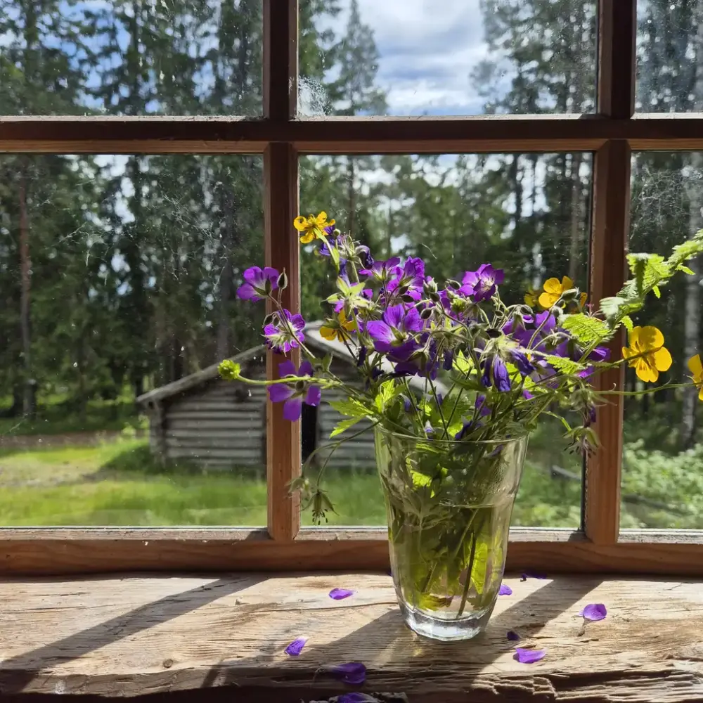 Ett glas med blommor i olika färger står i ett fönster med en låg byggnad i en skog utanför.