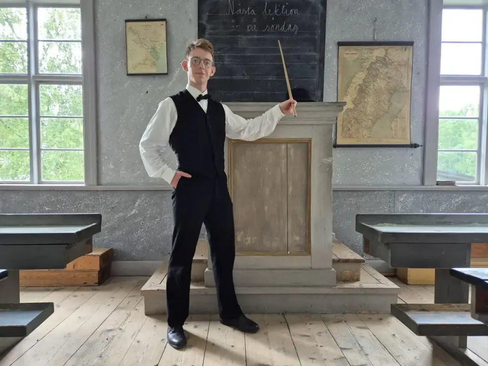 En person står i en skolsal och pekar med en pekpinne mot svarta tavlan.
