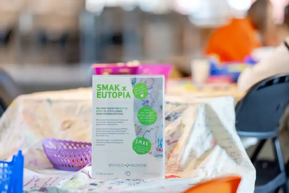 Plakat med informasjon om prosjektet Smak x Eutopia på bord med hvit duk med påsydde broderier
