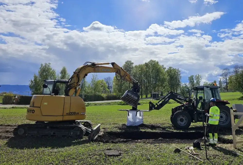 En gul gravemaskin fyller jord opp i en hvit storsekk som henger fast på pallegaffelen til en rød traktor. En arkeolog i singalgule klær står ved traktoren og følger med på arbeidet.