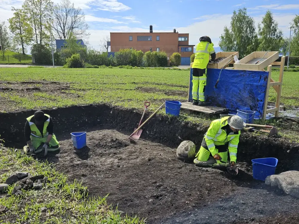 Et firkantet hull på ca. en halvmeters dybde er gravd i et jorde. Nede i hullet sitter to arkeologer i signalgule verneklær og graver med hver sin murerskje, med hver sin blå bøtte ved siden av. PÅ kanten bak hullet står en tredje arkeolog i gule verneklær og solder jord gjennom en svær sil bygget av planker og netting, solden står på skrå i en ramme.