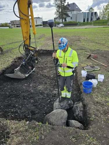 En arkeolog med signalgule klær og blå hjelm står ved noen store steiner nede i et firkantet hull som er gravd ut med gravemaskin. Grabben og deler av den gule armen på gravemaskinen vises, og bak arkeologen står flere plastbøtter klare.