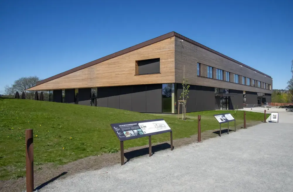 Anno Domkirkeodden historiesenters hovedbygning som åpnes 28. mai 2026 ser ut som en trekant av treverk som hviler på fire meter høye vindusfasader rundt det hele. På bildet ses byggets ene hjørne, med den høye fasaden til hovedinngangen på ene sida, og skråtaket ned mot restaurantens uteservering på andre sida. Bygget passer godt i landskapet.