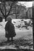 Ett okänt barn med mössa och vantar står på en snöig väg nedanför en fastighet.