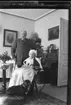 Porträtt av okänd äldre kvinna och man i hemmiljö. Mannen står med käpp bakom den sittande kvinnansom sitter på en stol i ljus tunn mössa och broderat ljust förkläde. Vid hennes fötter sitter det en tax.