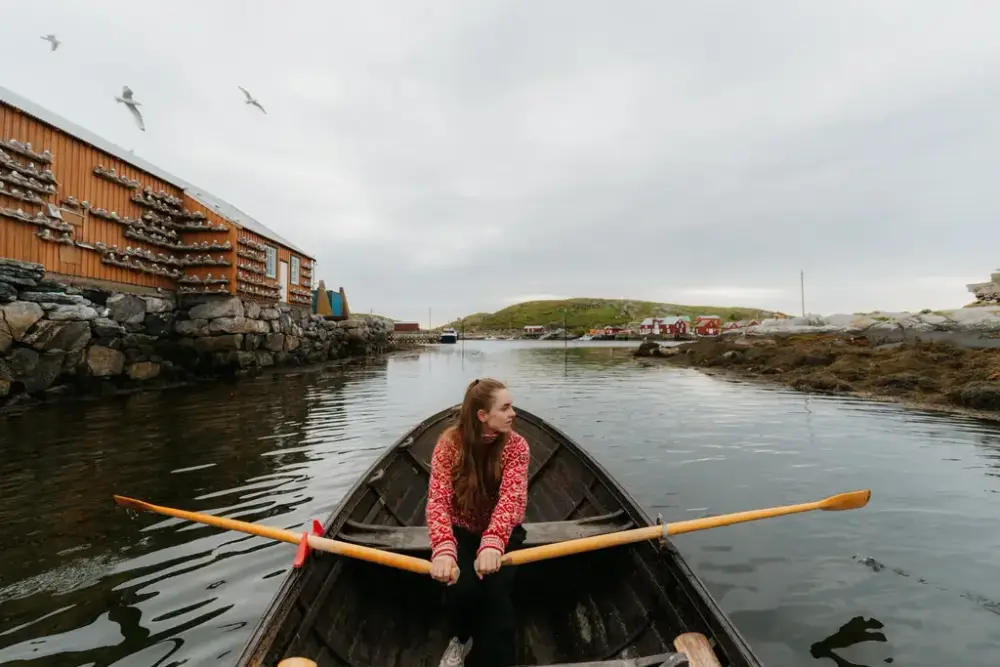 Kvinne i rød og hvit genser ror på vannet mellom et okergult hus på den ene siden og en holme på den andre siden. Foto.