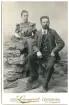 Familjefotografi över fabrikör M. F. R. Klinge med sin dotter Nora poserandes i en fotoatelje dekorerad med ett staket och stenhög.