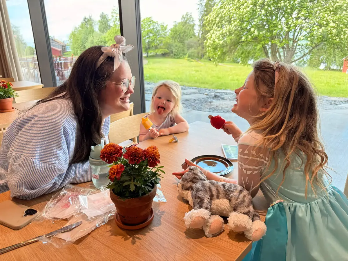 En mor og to barn sitter ved et kafébord med røde blomster og en kosebamse. Store vinduer viser grønn sommervegetasjon utenfor kafeen på Anno Domkirkeodden historiesenter.