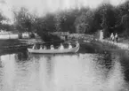 Sju personer i en roddbåt vid kanalen, Gröndal, Norrlandet, Gävle kommun 1902. Tre personer står på stranden.