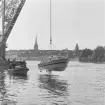 Motorbarkass sjösätts med kran vid Stockholms örlogsvarv. Skeppsbron och Gamla stan i bakgrunden.