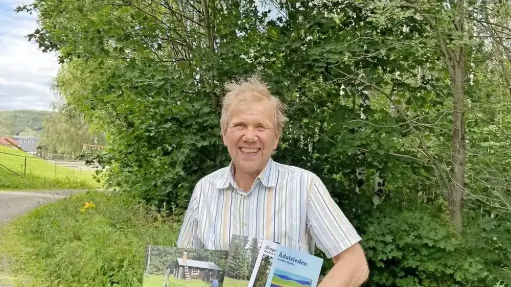 En man med ljus skjorta står framför grönskande träd med böcker i famnen.
