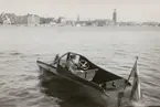 Mindre passbåt med inombordsmotor, på Riddarfjärden. Vy mot Norr Mälarstrand från Söder Mälarstrand. Namn på akterspegeln 