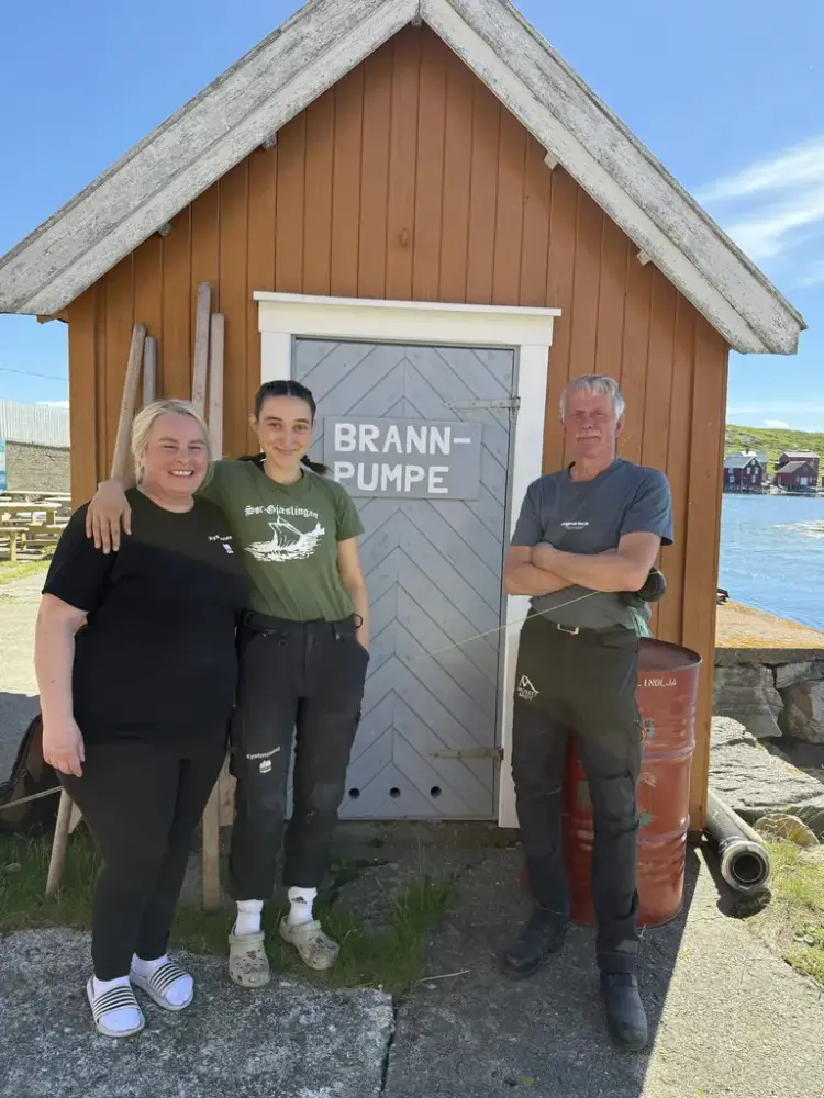 To kvinnelige ansatte og en mannligansatt står smilende foran et gult trebygg i solskinn, med vannet synlig i bakgrunnen. Foto.