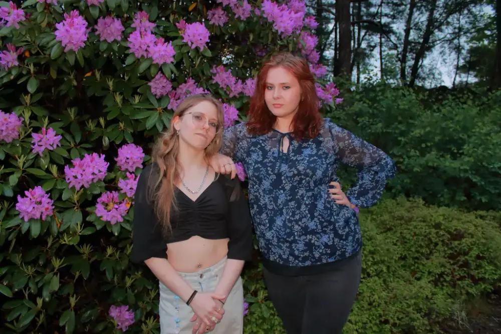To unge kvinner poserer i ein hage med kraftige lilla rhodondentronblomser