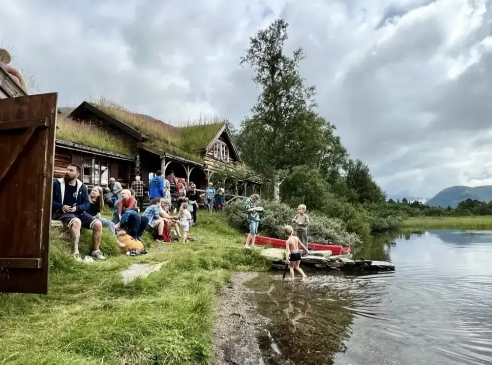 Mennesker ved et fjellvann med en gammel hytte bak