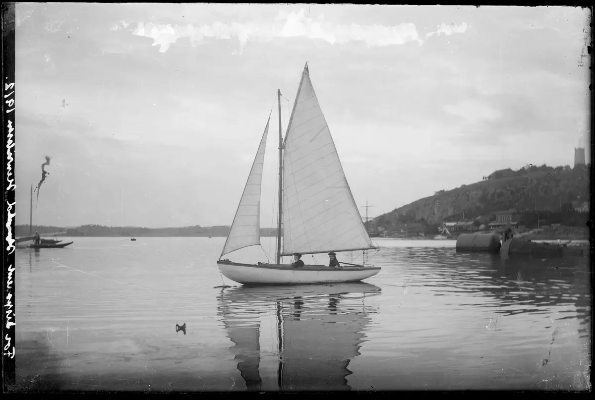 Sort-hvit bilde som viser en hvit seilbåt i en fjord. Øverst på toppen av skråningen 