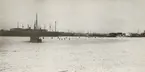 Isbelagd oljehamn, många cisterner, en infrusen båt och många människor på isen. Möjligen Värtahamnen.
