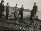 Män vid vatten, strand med byggnader i bakgrunden. Vedupplag och en båt vid brygga.