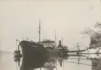 Fartyget Svithiod vid oljehamn, cisterner på land. Bogserbåten Frigga invid fartyget. Stämpel 