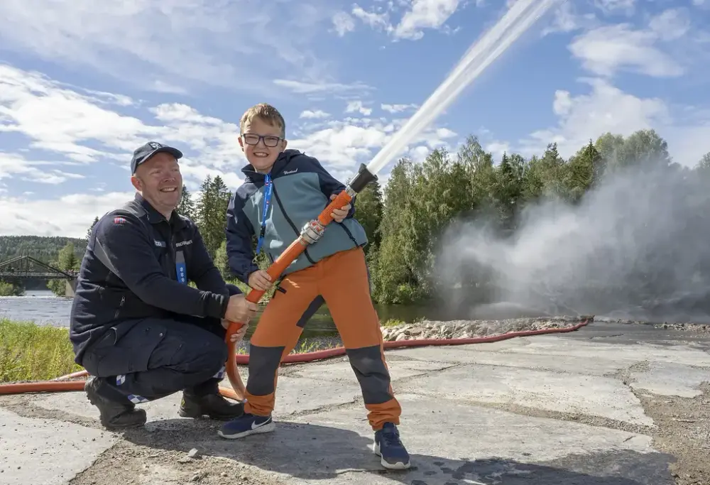 To personer i verneklær sprøyter vann med en brannslange utendørs under De nordiske jakt- og fiskedagene på Elverumsutstillingen.