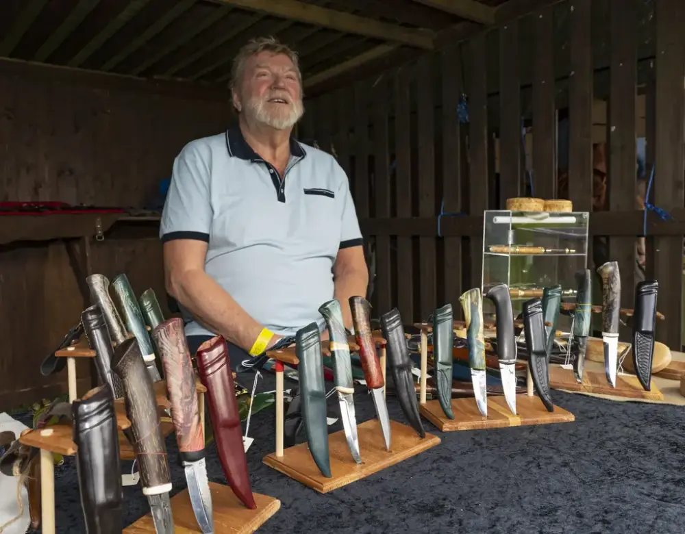 En mann bak en utstilling av håndlagde kniver på trestativer under De nordiske jakt- og fiskedagene på Elverumsutstillingen.