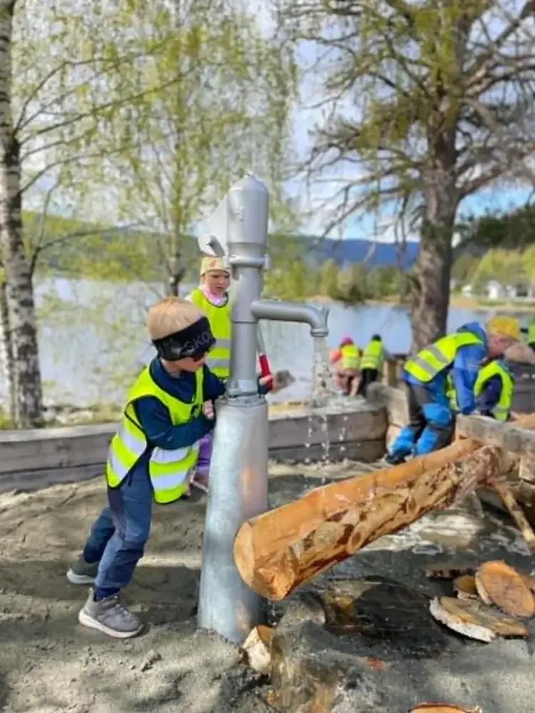 Barnehagebarn med refleksvestar som leiker med vasspumpe og vassrøyr i tre