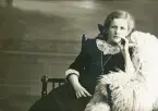 En flicka i mörk klänning och spetskrage sitter i en s.k. munkstol hos fotografen. 
Trol. Ruth Svensson (1901-1991), Alvesta, senare Stockholm.
