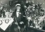 En student i smoking och studentmössa, avfotograferad i ett vardagsrum, omgiven av blommor.
Seved August Svensson (1894-1962), senare postassistent och kamrer, Skurup.