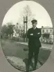 En ung man i mörk kavaj, knäbyxor och skolmössa poserar vid en gata (möjl. husen Ericsberg och Fridsberg vid Kungsvägen, Växjö).