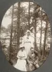 Två kvinnor i ljusa sommarklänningar och en man i kostym har slagit sig ner på en sten i skogen. I bakgrunden skymtar en sjö. 
Möjl. fru Helga Svensson med dotter Ruth och en okänd man.