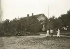 Tre personer framför gaveln till Östra folkskolan i Alvesta, folkskollärare Amos Svenssons hem under många år. I bakgrunden skymtar Aringsås (Alvesta) kyrka.
Längst till höger Ruth Svensson.