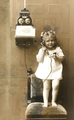 Liten jente står på stol og snakker i fasttelefon på veggen.