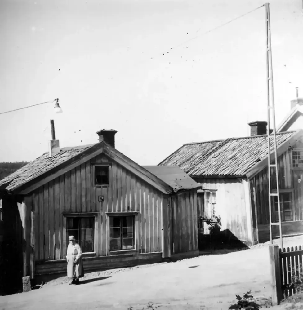 Gammalt foto av två små trähus på Långgatan 17A och B i Härnösand, en äldre kvinna har stannat upp och tittar på fotografen.