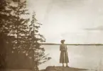 En ung kvinna i kappa och hatt (Ruth Svensson?) poserar på en bergsknalle vid en sjö.