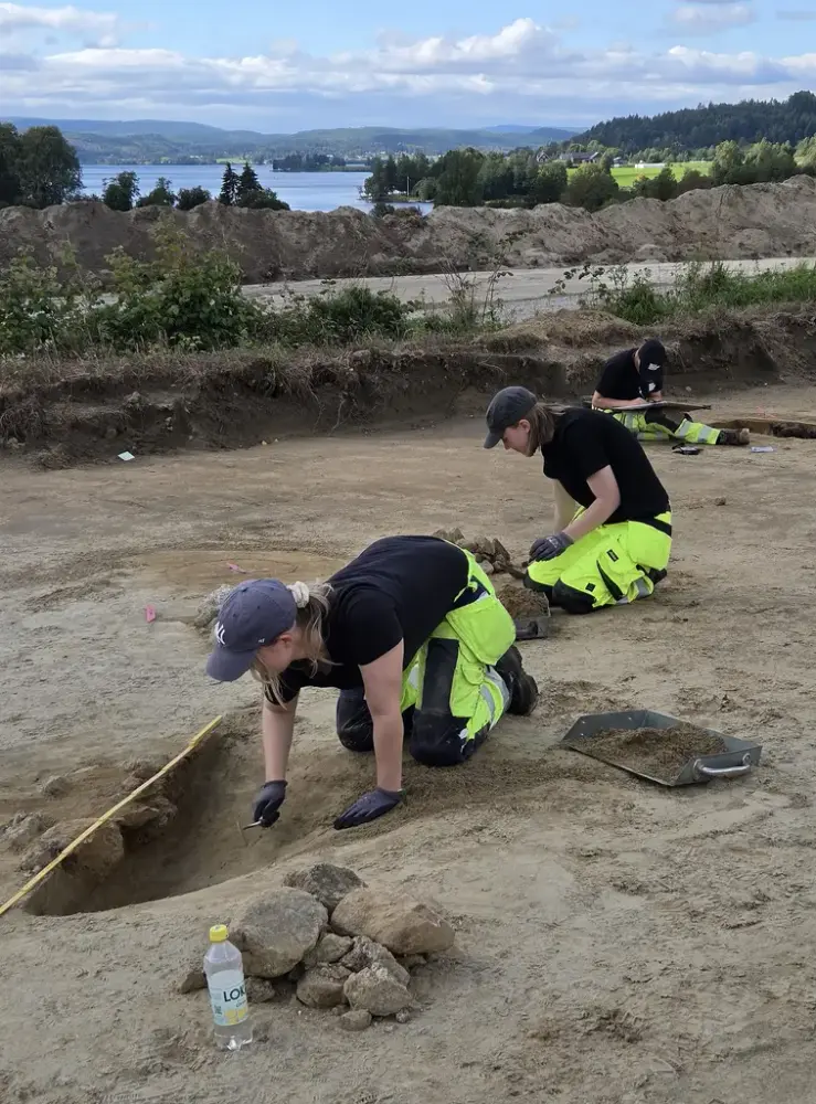 Två personer med keps, mörk tröja och varselbyxor gräver i uppgrävd jord.