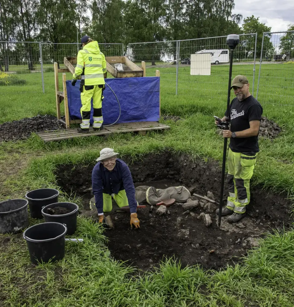 Et rektangulært hull ca. 70 cm dypt er gravd på en grønn gresslette. Til venstre for hullet står fire svarte plastbøtter, nede i hullet sitter arkeolog kjetil Skare på kne og graver, med en kollega stående til høyre i hullet. Bak hullet er det rigget en soldestasjon, og der står en arkeolog og spyler vann over jord som er tatt ut.