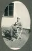 En man i ljus sommarkostym och studentmössa sitter på en trädgårdsstol på gårdpslanen till ett reveterat bostadshus. 
Något suddigt foto.