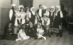 En grupp unga kvinnor och män i diverse olika folkdräkter och maskeraddräkter. I mitten sitter en man i mörk kostym och stärkkrage.
