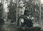 Två kvinnor och en flicka i promenaddräkt m.m. står på ett flyttblock i skogen.