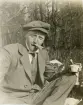 En man i kostym och keps sitter och dricker kaffe utomhus en solig vårdag. 
I munnen har han en stadig smörgås.
Trol. Yngve Svensson, bror t. Ruth.