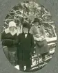 Två kvinnor i kappa och hatt och en man i rock med sammetskrage och pälsmössa (persian) har stannat upp vid en gärdesgård,
under en vintrig promenad.