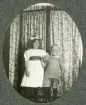 Två små barn, en flicka och en pojke, poserar framför en skärm med småblommigt tyg. 
Något suddigt foto.