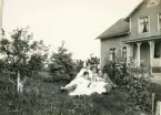 Fyra kvinnor i ljusa sommarklänningar och två män i kostym m.m. har slagit sig ner på en gräsmatta i en trädgård. I bakgrunden syns ett bostadshus med veranda.