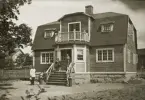 Ett nybyggt bostadshus med trädgård. På trappan framför huset syns en medelålders man och en kvinna. Till vänster kommer en man via gårdsplanen med leverans av en tidning m.m.
Kalmar (?).