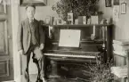 En man i kostym med stärkkrage och ljus väst poserar vid ett piano. 
Pianot verkar vara levererat av firman 