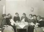En syförening (?). Sju kvinnor med diverse handarbeten har samlats kring ett bord.